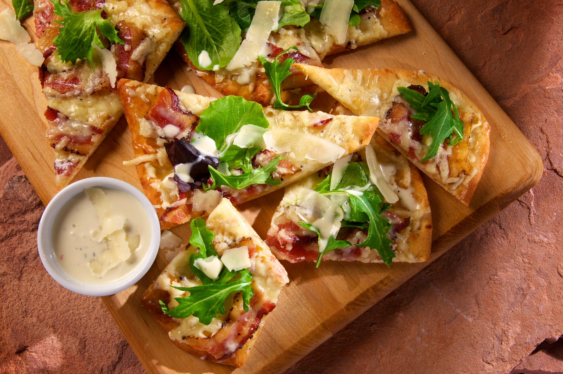 This unique appetizer pizza–and great party food idea–features thin crispy flatbread baked golden and delicious topped with crisp peppered bacon and gooey melted Italian cheese. It’s finished with a drizzle of indulgent garlic Parmesan cream sauce, fresh spring greens and shaved Parmesan cheese.