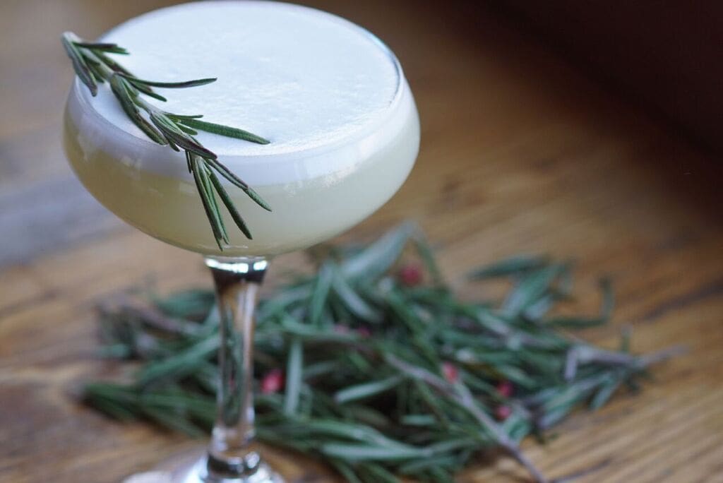 Add egg white and gin in a Boston shaker and shake 5-6 times. Add in liqueur, lemon juice, rosemary syrup, and olive oil, then fill shaker with ice. Shake vigorously and strain in to a cocktail glass. Garnish with bitters and Rosemary sprig.